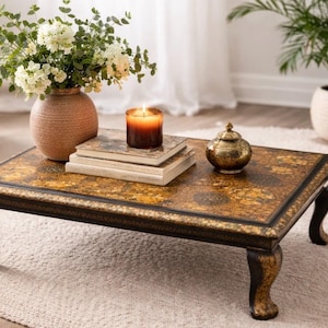 Puede incluir: Una mesa de centro rectangular con detalles dorados y negros ornamentados. La mesa tiene patas curvas y una tapa decorativa. Un jarrón con flores blancas, libros, una vela y un tarro dorado están sobre la mesa.