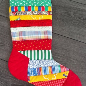 May include: A quilted Christmas stocking constructed from horizontal fabric strips featuring a variety of patterns including floral, plaid, polka dot, and paisley prints in red, green, yellow, blue, and white, with a red quilted heel and toe.