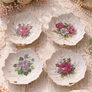 May include: Four white leaf-shaped porcelain dishes with gold trim and floral designs. Each dish features a different arrangement of colorful flowers, including roses and violets. The dishes are arranged on a light-colored wooden surface.