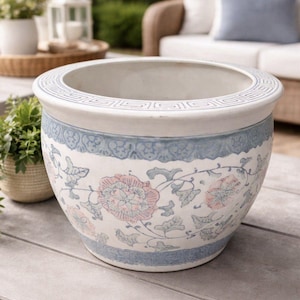 Puede incluir: Una maceta de cerámica grande y redonda con base blanca y diseños florales y geométricos azules. La maceta tiene una abertura ancha y un borde azul decorativo. Flores rosas y hojas verdes pintadas en la maceta.