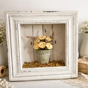 Op de afbeelding: Een vierkant, wit, verweerd schaduwboxframe met een kleine, verouderde groene pot gevuld met gele rozen. De pot staat op mos in het frame, waardoor een miniatuur bloemenarrangement ontstaat. Het frame heeft een rustieke, vintage uitstraling.