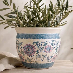 May include: A round ceramic planter with a cream-coloured base, featuring a decorative blue border at the top and bottom. The centre is adorned with a repeating pattern of pink and blue flowers and green leaves. An olive branch is inside the planter.