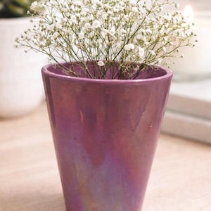 May include: A small, glossy, purple vase holds a bouquet of white baby's breath flowers. The vase has a pearlescent finish, reflecting light with hints of gold and pink. The flowers are delicate and airy, creating a soft contrast with the vase.