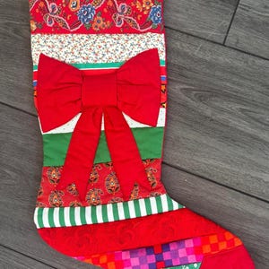 May include: A quilted Christmas stocking featuring a patchwork design of various fabric patterns including red floral, green stripes, and a large central red fabric bow. The stocking is constructed from horizontal bands of colorful textiles.