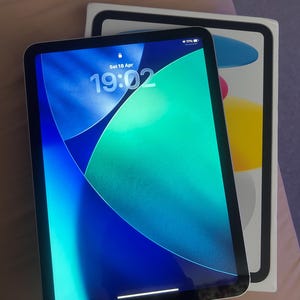 May include: A black tablet with a vibrant blue and green screen displaying the time and date. The tablet is next to its white box, which has a colorful design. The screen shows the time as 19:02.