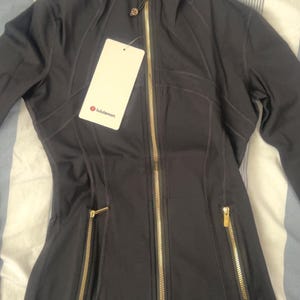 May include: Dark gray athletic jacket with a high collar and gold-colored zippers. The jacket has a fitted silhouette, two front pockets, and a tag with the brand name "lululemon".