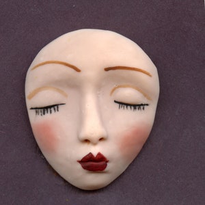 May include: A ceramic face with closed eyes, painted with light skin, red lips, and blush on the cheeks. The eyebrows are brown, and the eyelashes are black. The face is oval-shaped and appears to be a decorative item.