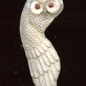 May include: A carved owl figurine in off-white, featuring detailed feather and scale textures. The owl has large, round, brown eyes and a curved body, giving it a unique, artistic appearance. The figurine is set against a dark background.