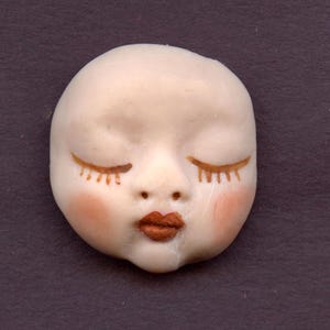 May include: A white porcelain doll face with closed eyes and brown eyelashes. The doll has rosy cheeks and a brown painted mouth.