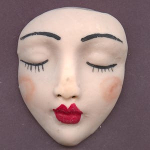May include: A ceramic face with closed eyes, black eyebrows, and long black eyelashes. The face has red lips and pink blush on the cheeks. The background is a solid, muted purple.
