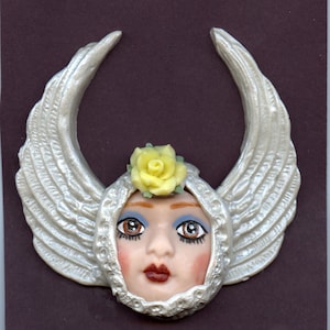 May include: A decorative angel face with silver wings and a yellow rose. The face has brown eyes, blue eyeshadow, and red lips. The wings are textured and surround the face, creating a unique, artistic design.