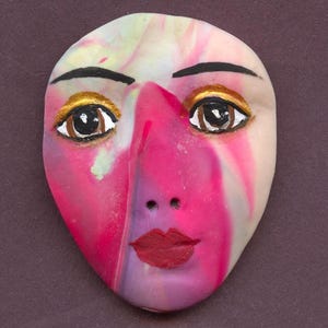 May include: A handcrafted mask with a face design. The mask features a pink and white marbled pattern, with painted details including brown eyes, gold eyeshadow, black eyebrows, and red lips. The mask is an oval shape.