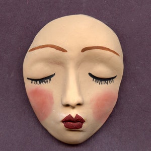 May include: A ceramic face mask with closed eyes, painted with brown eyebrows, black eyelashes, pink blush, and a red lip.