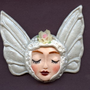 May include: A decorative fairy face with closed eyes, rosy cheeks, and dark red lips. The fairy has large, silver-colored wings and a small pink rose on her head. The face is framed by a silver-colored hood with intricate detailing.