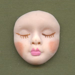 May include: A small, pale-skinned face with closed eyes, painted eyelashes, and pink blush on the cheeks. The lips are painted a bright pink. The face is set against a green background.