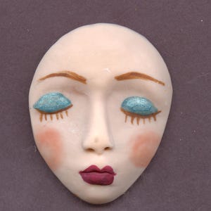 May include: A ceramic face with closed eyes, painted with light skin tone, brown eyebrows, and teal eyeshadow. The cheeks are blushed pink, and the lips are a deep burgundy. The face is oval-shaped and has a smooth, matte finish.