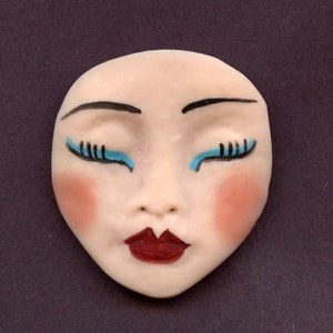 May include: A ceramic face with closed eyes, painted with black eyebrows and eyelashes, blue eyeliner, red lips, and pink blush on the cheeks. The face is a light beige color and has a smooth finish.