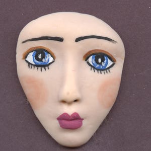 Op de afbeelding: Een handgeschilderd keramisch gezicht met blauwe ogen, zwarte wenkbrauwen en roze blos. De lippen zijn diep magenta geschilderd. Het gezicht heeft een gladde, bleke teint en is waarschijnlijk een decoratief item of onderdeel van een groter kunstwerk.