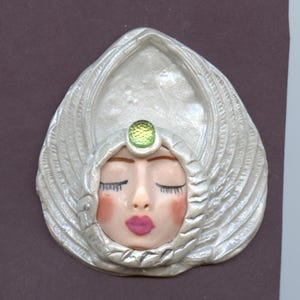 May include: A white ceramic sculpture of a woman's face with closed eyes. The woman is wearing a silver headdress with a green gem in the center.