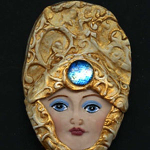 May include: A decorative face sculpture with a golden headdress and a blue gemstone. The face has blue eyes, light skin, and red lips. The headdress has an ornate, textured design.