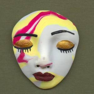 May include: A ceramic face mask with closed eyes, painted with yellow, pink, and white hues. The eyelids are gold, with black eyelashes and eyebrows. The lips are a deep burgundy color, and the mask has a smooth, matte finish.