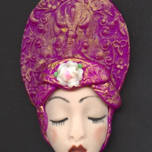 May include: A decorative face sculpture with closed eyes, featuring a magenta headdress with gold accents and a white rose. The face has a fair complexion, rosy cheeks, and dark red lips. The sculpture is set against a black background.