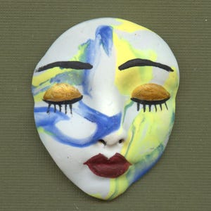 May include: A ceramic face mask with closed eyes, painted with blue, yellow, and white swirls. The eyelids are gold, the eyelashes are black, and the lips are a deep red. The mask has a smooth, matte finish.