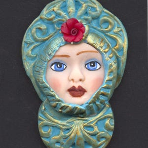May include: A decorative face sculpture with a turquoise and gold color scheme. The face has blue eyes, rosy cheeks, and a red rose on the head. The sculpture is an oval shape with ornate detailing.