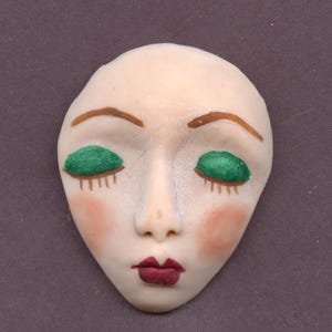 May include: A ceramic face with closed eyes, painted with green eyeshadow, brown eyebrows, and red lips and cheeks. The face is oval-shaped and has a smooth, pale complexion. The background is a solid, muted purple.