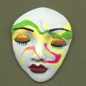 May include: A white ceramic mask with closed eyes and a painted face. The mask features gold-colored eyelids, black eyelashes, and a red mouth. Swirls of yellow, green, and pink paint decorate the face, creating a unique artistic design.