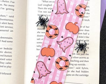 May include: A pink and white gingham bookmark with a Halloween theme. The bookmark features a repeating pattern of ghosts, pumpkins, and donuts with bats. There are three black plastic spiders on the bookmark.