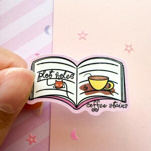 May include: A sticker featuring an open book design with the words "plot holes" and "coffee stains." The book illustration includes a cartoon cat and a yellow coffee cup. The sticker is held by a hand against a pink background.