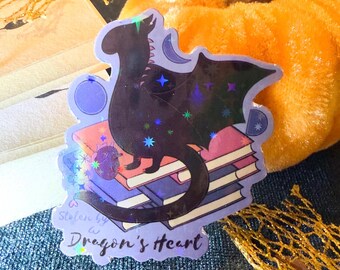 May include: A holographic sticker featuring a black dragon with purple wings perched on a stack of books. The sticker has a blue background with stars and a crescent moon. The text "Stolen by a Dragon's Heart" is printed on the sticker.