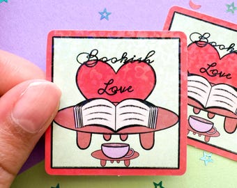 May include: Square sticker with a red heart and the words "Bookish Love" in cursive. An open book is below the heart, with a small teacup at the bottom. The sticker has a red border and is on a light green background.