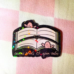 May include: A holographic sticker featuring an open book with two cacti peeking out from the pages. The text "cactus trails & open tales" is printed below the book.