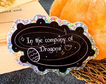 May include: A black and white holographic sticker with a glittery finish. The sticker features a black oval with the text "In the company of Dragons" in a white, cursive font. The oval is surrounded by a white, glittery border.