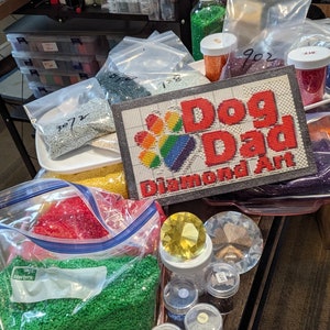 May include: A close-up of a variety of colorful diamond painting kits, including a kit with a rainbow paw print and the text "Dog Dad Diamond Art".