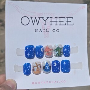 May include: A set of ten press-on nails on a white display card labeled OWYHEE NAIL CO. The nails feature a blue night sky theme with white stars, moons, a cartoon hippopotamus, a green alligator, a brown broom, and a blue wizard hat design.