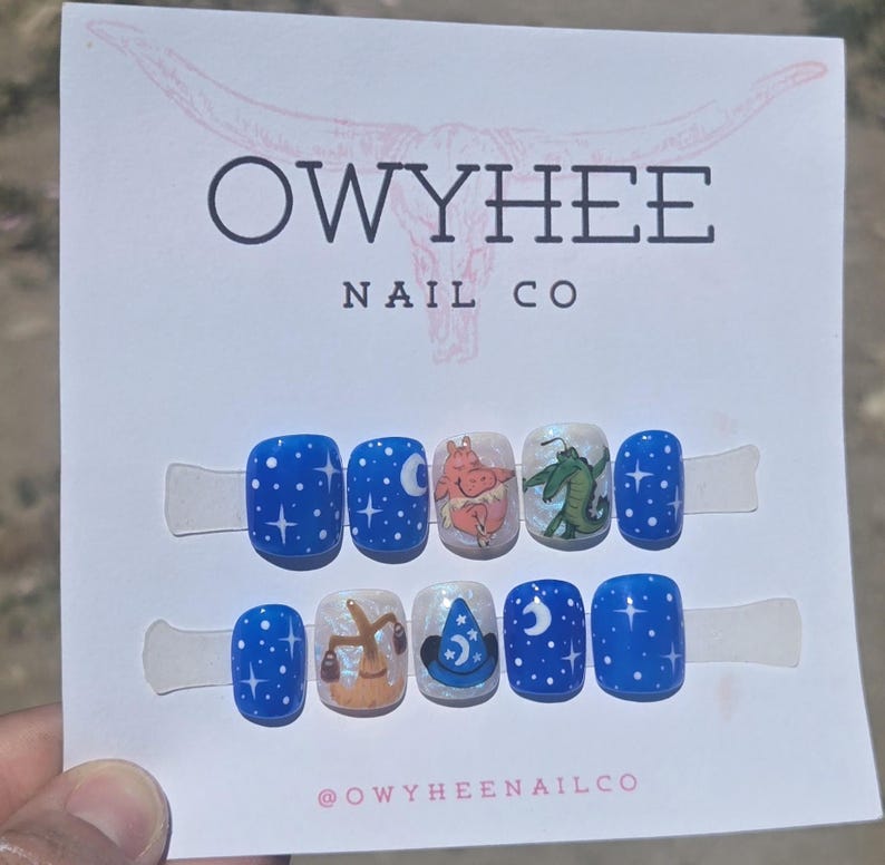 May include: A set of ten press-on nails on a white display card labeled OWYHEE NAIL CO. The nails feature a blue night sky theme with white stars, moons, a cartoon hippopotamus, a green alligator, a brown broom, and a blue wizard hat design.