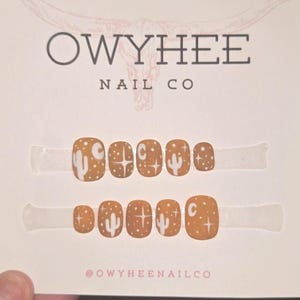 May include: A set of ten press-on nails in a matte mustard yellow finish, featuring white hand-painted designs of cacti, stars, and crescent moons. The nails are displayed on a white card labeled Owyhee Nail Co with a bull skull graphic.