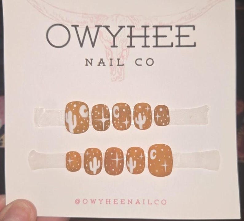 May include: A set of ten press-on nails in a matte mustard yellow finish, featuring white hand-painted designs of cacti, stars, and crescent moons. The nails are displayed on a white card labeled Owyhee Nail Co with a bull skull graphic.