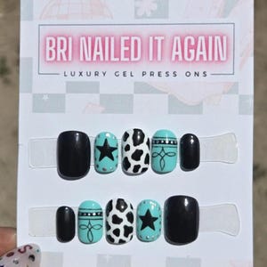 May include: A set of ten press-on nails on a display card labeled BRI NAILED IT AGAIN LUXURY GEL PRESS ONS. The nails feature black, white, and turquoise designs including cow print patterns, black stars, and decorative line work with silver dots.