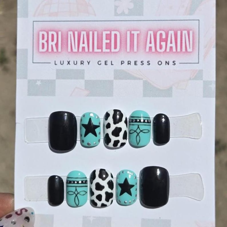 May include: A set of ten press-on nails on a display card labeled BRI NAILED IT AGAIN LUXURY GEL PRESS ONS. The nails feature black, white, and turquoise designs including cow print patterns, black stars, and decorative line work with silver dots.