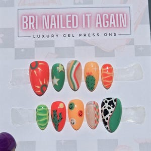 May include: A set of ten almond-shaped press-on nails featuring vibrant designs including cacti, smiley faces, cow print, suns, and stars in shades of orange, green, and white. The display card reads BRI NAILED IT AGAIN LUXURY GEL PRESS ONS.