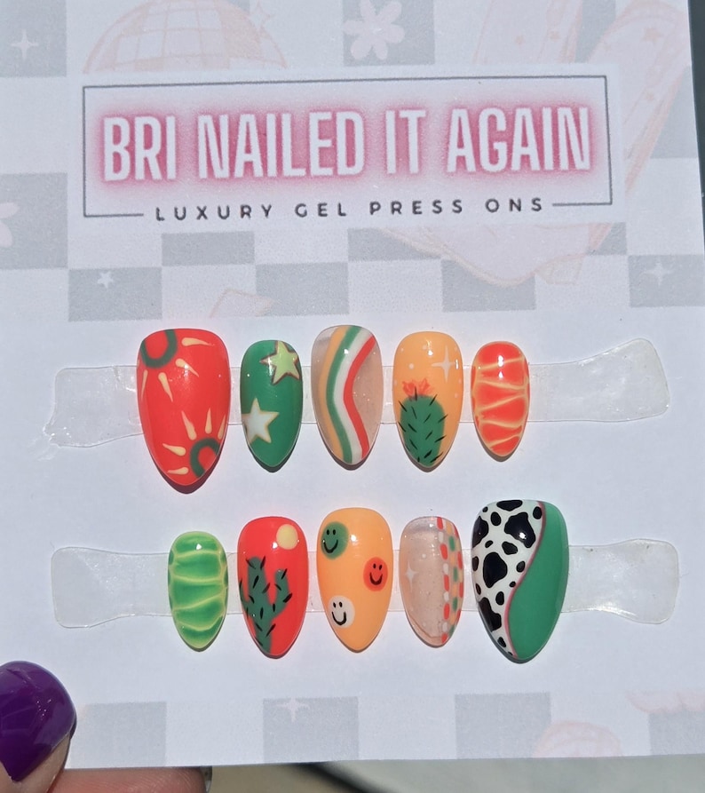 May include: A set of ten almond-shaped press-on nails featuring vibrant designs including cacti, smiley faces, cow print, suns, and stars in shades of orange, green, and white. The display card reads BRI NAILED IT AGAIN LUXURY GEL PRESS ONS.