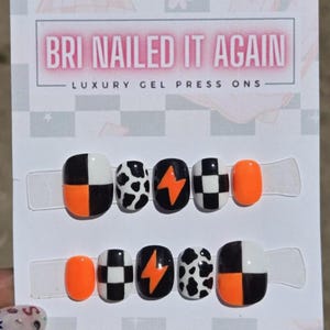 May include: A set of ten press-on gel nails on a display card labeled BRI NAILED IT AGAIN LUXURY GEL PRESS ONS. The nails feature black, white, and neon orange designs including checkerboard patterns, cow print, and orange lightning bolts.