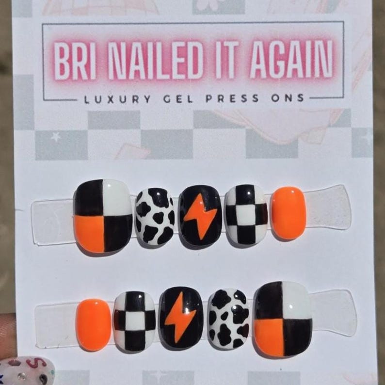 May include: A set of ten press-on gel nails on a display card labeled BRI NAILED IT AGAIN LUXURY GEL PRESS ONS. The nails feature black, white, and neon orange designs including checkerboard patterns, cow print, and orange lightning bolts.