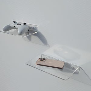 May include: Two clear acrylic wall shelves. The top shelf holds a white and gray game controller. The bottom shelf holds a gold-colored smartphone. Both shelves are mounted on a white wall.