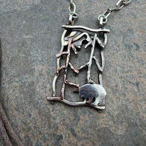 May include: Silver-tone pendant necklace featuring a bear silhouette within a rectangular frame. The frame has a forest-like design with tree and leaf details. The necklace is on a gray surface.