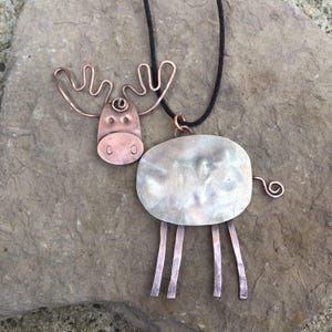 May include: A handcrafted copper moose pendant necklace. The pendant features a stylized moose design with a copper head, antlers, and legs, and a silver-toned body. The necklace has a black cord.